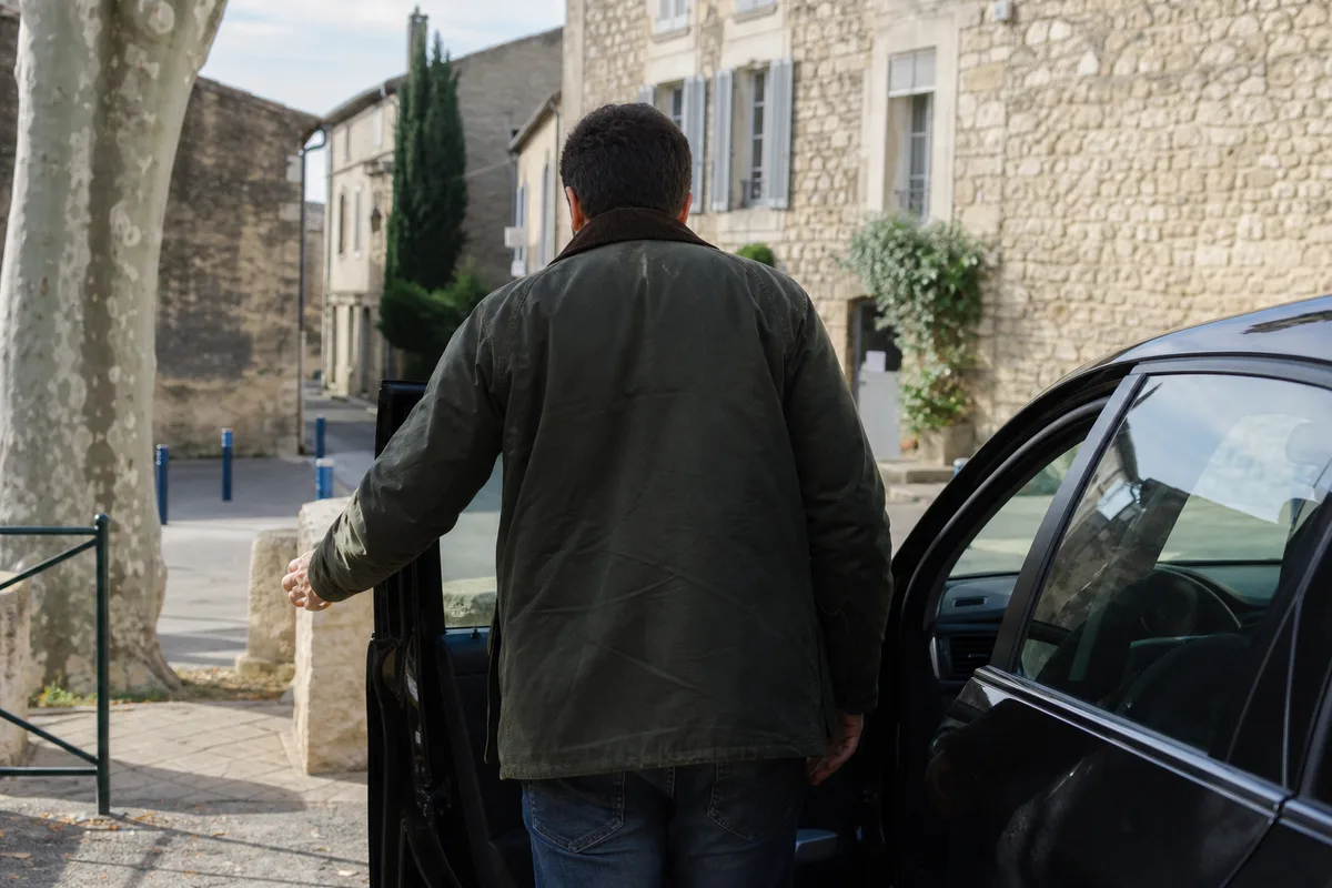 Silhouette de dos d&rsquo;une personne ouvrant la portière d&rsquo;une voiture sur une place de village française aux façades en pierre