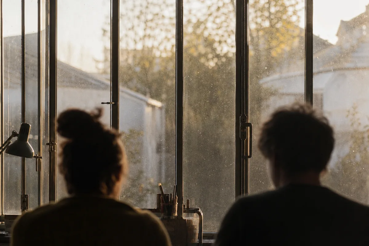 Silhouettes de deux personnes assises de dos près d&rsquo;une grande fenêtre d&rsquo;atelier au petit matin