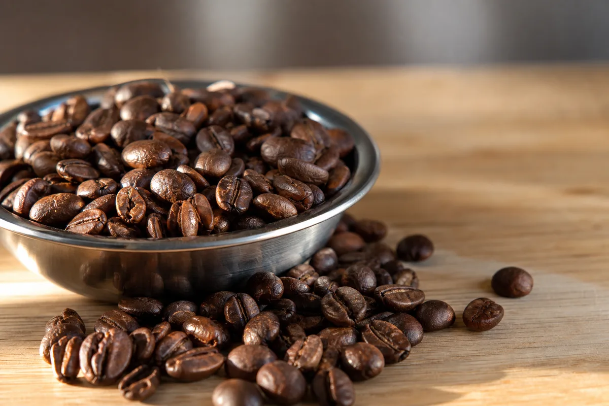 Grains de café torréfiés en vrac sur une surface en bois clair, lumière rasante de fin de journée