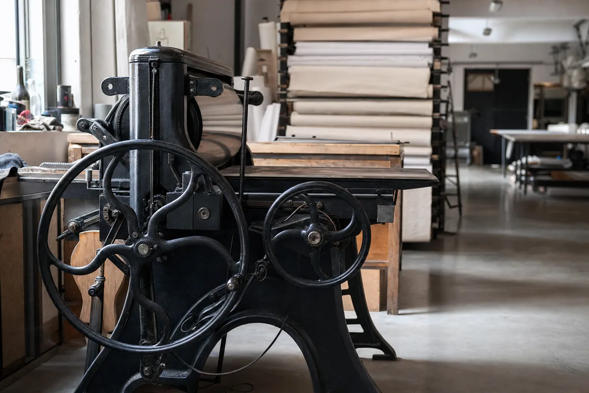 Atelier d&rsquo;imprimerie française baigné de lumière naturelle, presse ancienne et rouleaux de papier en pile